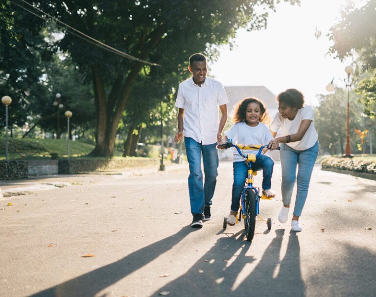 A black family teaching their young child to ride a bike in a park, representing the intergenerational support needed to build generational wealth.