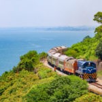 Train traveling on track with blue sky, ocean water and green trees.
