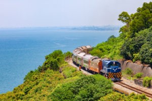 Train traveling on railroad with blue sky, ocean water and green trees.