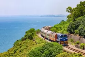 Train traveling on track with blue sky, ocean water and green trees.