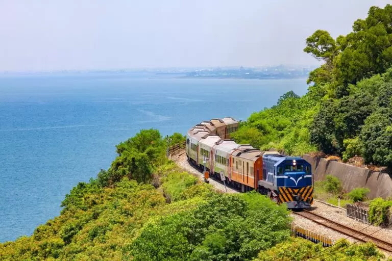 Train traveling on track with blue sky, ocean water and green trees.