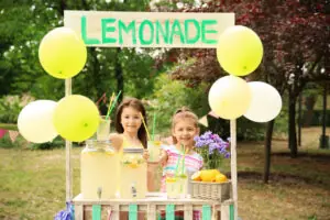 Two kids smiling behind a wooden lemonade stand outside, holding up a glass of lemonade for a customer.