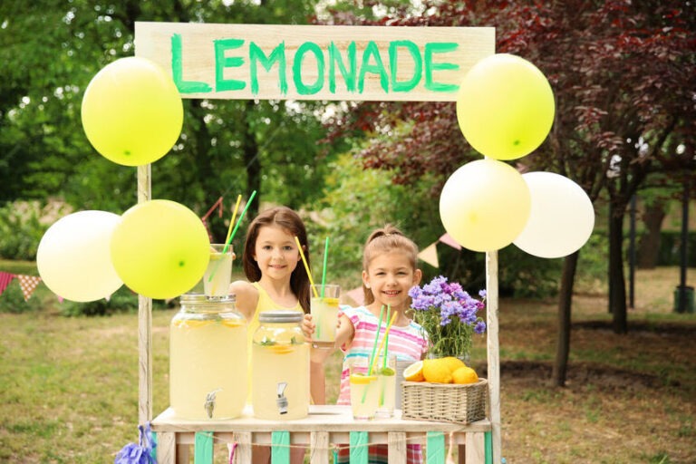 Two kids smiling behind a wooden lemonade stand outside, holding up a glass of lemonade for a customer.