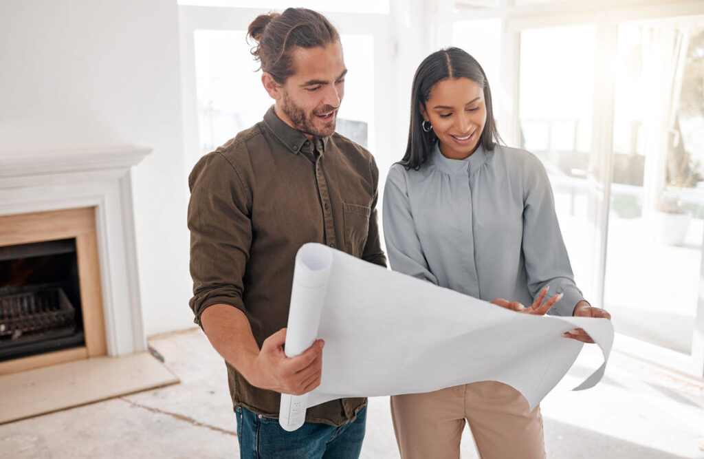 Reviewing Home Renovation Loan Project A diverse couple stands in the living room reviewing architectural blueprints and plans for their home renovation project.